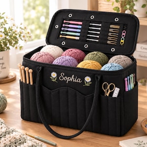 May include: A black rectangular yarn storage bag with the name Sophia embroidered in white between two daisy flowers. The open lid reveals slots for crochet hooks and needles. The exterior features pockets holding wooden needles, scissors, and hooks.