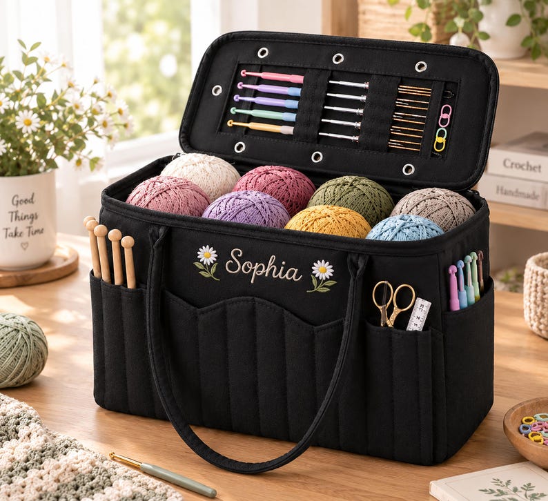 May include: A black rectangular yarn storage bag with the name Sophia embroidered in white between two daisy flowers. The open lid reveals slots for crochet hooks and needles. The exterior features pockets holding wooden needles, scissors, and hooks.