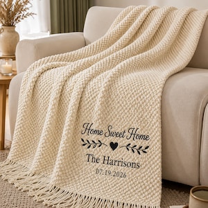 May include: A cream-coloured chunky knit throw blanket with fringe edges. The blanket features black embroidered text that reads Home Sweet Home, The Harrisons, and the date 07.19.2026, accented by a central heart and two leafy branch motifs.