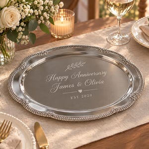 May include: An oval silver-coloured serving tray with a decorative beaded and floral embossed rim. The centre is engraved with a leaf sprig, the text "Happy Anniversary James & Olivia", a small heart icon, and the text "EST 2020" in a clean font.