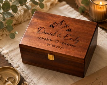 May include: A rectangular dark wood keepsake box with a hinged lid and a gold-tone metal latch. The lid features a laser-engraved mountain range, the names Daniel & Emily, a small heart, leafy branches, and the date 05.18.2026 in black ink.
