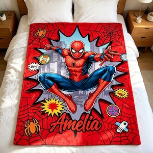 May include: A red fleece blanket featuring a graphic of a superhero in a red and blue suit leaping against a city skyline background. The design includes black spiderweb patterns, colorful comic-style bursts, and the name Amelia in black script.