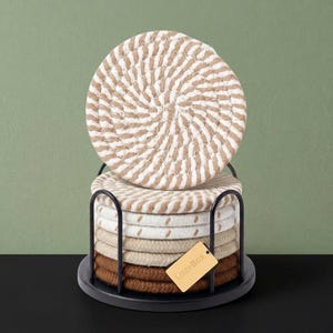 May include: A stack of six round, woven rope coasters in shades of beige, cream, and brown, held in a black metal wire stand. One coaster stands vertically on top, while the others are stacked horizontally. A small tan tag reads LazeBox.