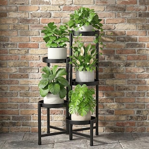 May include: A black metal tiered plant stand with five circular shelves arranged at varying heights. The stand holds five potted plants, including a peace lily with white blooms and a monstera, in white textured ceramic pots of different designs.