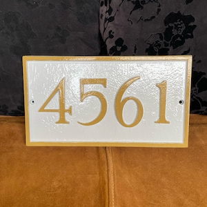 以下が含まれることがあります： 茶色の表面に、金色の縁取りと金色の数字「4561」がついた白い家の番号プレート