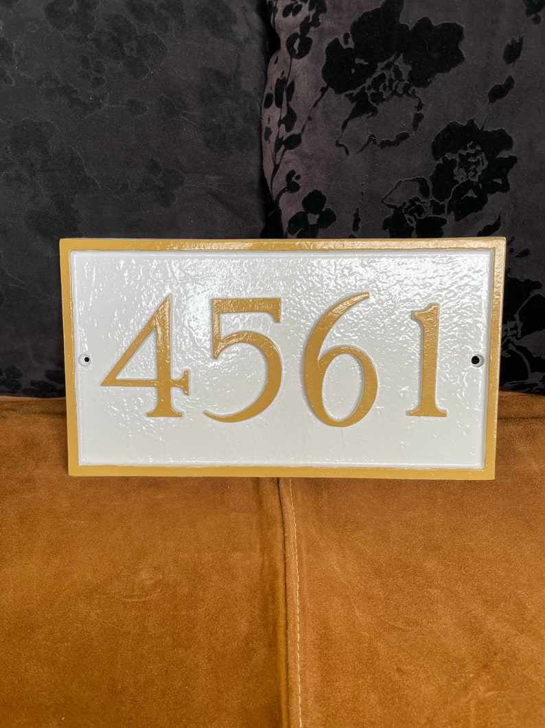 以下が含まれることがあります： 茶色の表面に、金色の縁取りと金色の数字「4561」がついた白い家の番号プレート