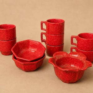 May include: A collection of red ceramic tableware featuring a mottled finish. The set includes six cylindrical mugs with square handles, two octagonal bowls, and one bowl shaped like a flower with scalloped edges, all sharing a consistent red glaze.