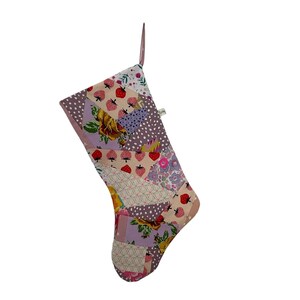 May include: A patchwork Christmas stocking with a variety of fabric patterns. The patterns include strawberries, floral designs, and polka dots in shades of pink, purple, and white. A pink loop is attached for hanging.