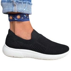 May include: Black slip-on sneakers with a white sole and a knit upper. The sneakers are worn with blue jeans and a blue and orange ankle bracelet with a celestial pattern.
