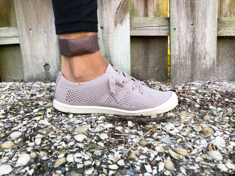 May include: A close-up of a person's ankle wearing a brown ankle strap. The person is wearing a pair of light grey knitted trainers with white soles.