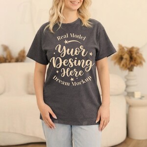 May include: A dark heather grey short-sleeve t-shirt featuring cream-coloured text that reads Real Model Yuor Desing Here Dream Mockup, arranged in a circular pattern with small star accents. The shirt has a crew neckline and a standard fit.