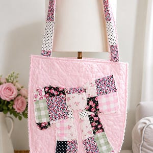 Op de afbeelding: Een doorgestikte lichtroze tote bag met een grote, centrale patchwork strik. De strik is gemaakt van diverse stoffen vierkantjes met bloemen-, ruitjes- en stippenpatronen. De tas heeft twee bijpassende doorgestikte schouderbanden.