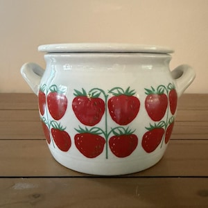 Peut inclure: Un pot en céramique blanche avec un couvercle et deux anses latérales, orné d'un motif répétitif de fraises rouges avec des tiges et des feuilles vertes disposées sur deux rangées horizontales autour du récipient rond.