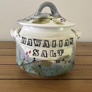 May include: A ceramic jar with a lid and two side handles, featuring a white base with blue, green, and red painted botanical accents. The text "HAWAIIAN SALT" is embossed in black lettering on the front of the jar, which has a speckled finish.