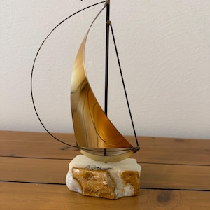 Könnte beinhalten: Eine Segelboot-Skulptur aus Metall mit einem polierten, goldfarbenen, geschwungenen Segel und einem dünnen, dunklen Mast mit einer kleinen Kupferflagge an der Spitze. Das Boot steht auf einem rauen, rechteckigen weiß-beigen Steinsockel mit Signatur.