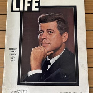 Pode incluir: Uma capa de revista LIFE vintage de 29 de novembro de 1963, com um retrato a cores do presidente John F. Kennedy com a mão sob o queixo. O texto diz President John F. Kennedy, 1917-1963, e inclui uma etiqueta de envio na parte inferior.