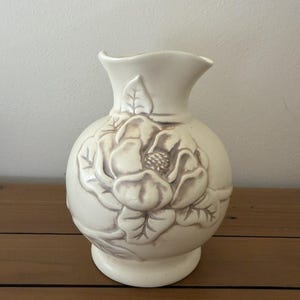 May include: A cream-colored ceramic vase with a rounded body and a flared rim. The exterior features a raised, embossed floral design with light brown detailing on the petals and leaves, creating a textured appearance on the smooth surface.