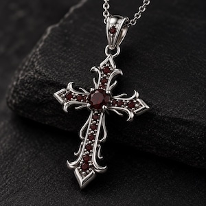 May include: A silver-toned metal cross pendant suspended from a matching chain. The cross features intricate scrollwork detailing and is set with multiple small, round, deep red gemstones, including a larger central stone and one on the bail.