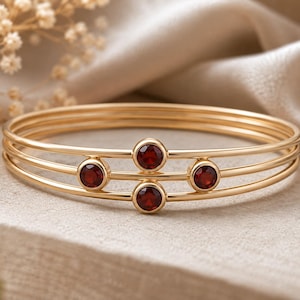 Peut inclure: Un bracelet jonc doré composé de trois anneaux fins superposés, reliés par quatre pierres précieuses grenat rouge foncé, serties clos et disposées en un motif géométrique décalé sur le devant de la structure métallique circulaire.