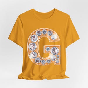 Può includere: Una t-shirt a maniche corte color giallo senape con una grande grafica centrale della lettera G. La lettera è resa in una tonalità oro metallizzato ed è impreziosita da numerosi cristalli bianchi circolari sfaccettati che imitano i diamanti.
