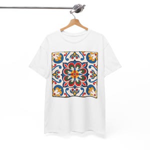 Può includere: Una t-shirt bianca a maniche corte con una stampa grafica quadrata centrata. Il design è costituito da un motivo simmetrico e complesso nei toni del blu, rosso, verde e giallo, che ricorda una piastrella decorativa con motivi floreali.