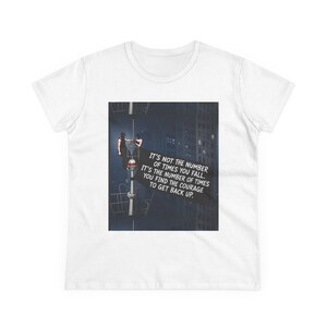 May include: A white short-sleeved t-shirt featuring a graphic print of two figures on a tall structure at night. The text reads: IT'S NOT THE NUMBER OF TIMES YOU FALL. IT'S THE NUMBER OF TIMES YOU FIND THE COURAGE TO GET BACK UP. The design is centered.