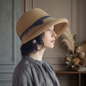 May include: A tan straw hat with a wide, slightly downturned brim and a dark navy blue ribbon band tied in a small bow on the side. The hat features a textured, woven material construction and a rounded crown, providing a classic look.