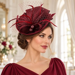Könnte beinhalten: Ein tiefburgunderroter Fascinator mit einer strukturierten Basis aus Stoffschlaufen, zarten Federn und einem feinen Netzschleier. Der Kopfschmuck ist mit zwei kleinen, texturierten kugelförmigen Akzenten in derselben dunkelroten Farbe verziert.