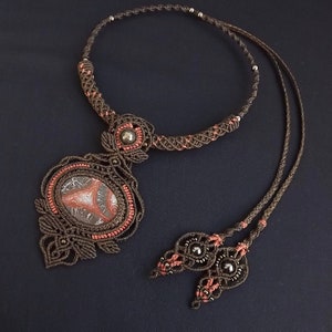 May include: A handcrafted macrame necklace featuring a large, oval-shaped stone pendant with brown, white, and red patterns. The necklace is made with brown cord and accented with small beads and coral-colored details. The adjustable cord is approximately 20 inches long.