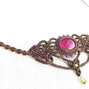 Può includere: Una collana in macramè marrone con un ciondolo in pietra preziosa rosa e perline viola. La collana è realizzata con una tecnica di annodatura e presenta un design delicato e intricato.