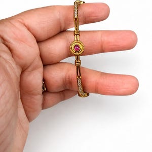 May include: A gold-toned bangle bracelet featuring a central circular medallion with a small pink stone. The band is accented with segments of clear, sparkling stones and decorative metal bars, creating a structured and detailed design.