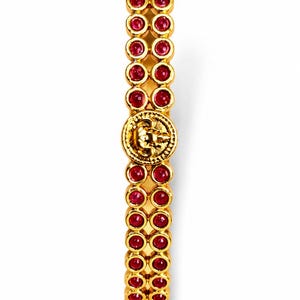 May include: A gold-toned bangle bracelet featuring two parallel rows of small, round, deep red stones. The bracelet is accented with a central circular gold medallion displaying an embossed design and decorative gold beadwork at both ends.