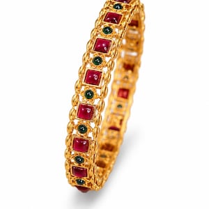 May include: A gold bangle bracelet featuring a repeating pattern of square-cut red stones and small round green stones. The metal band has a textured, filigree-style design with raised edges framing the inlaid gemstones in a continuous circular row.