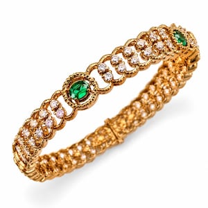 May include: A gold-toned hinged bangle bracelet featuring a repeating pattern of circular links. The design includes two oval-cut green stones set in textured gold frames, interspersed with rows of small, clear, round-cut white stones.