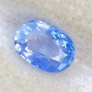Puede incluir: Una piedra preciosa azul de corte ovalado con una superficie facetada que refleja la luz. La piedra muestra un color azul aciano vibrante y translúcido con un acabado pulido y facetas geométricas simétricas en toda su superficie.