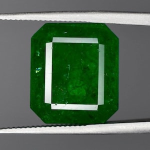Puede incluir: Una piedra preciosa de color verde vibrante con corte esmeralda, que presenta una forma rectangular con esquinas biseladas. La piedra muestra un tono verde profundo y saturado con inclusiones internas visibles y facetas que reflejan la luz.