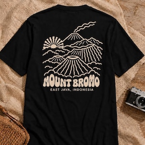 Peut inclure: Un t-shirt noir à manches courtes avec un motif graphique en lignes beige clair au dos. Le dessin représente le mont Bromo avec un soleil, des nuages et de la fumée, accompagné du texte MOUNT BROMO et EAST JAVA, INDONESIA en police arrondie.