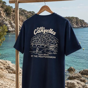 Può includere: Una t-shirt a maniche corte blu navy con una grafica a linee bianche sul retro. Il disegno raffigura un villaggio costiero, una persona su un paddleboard e il testo "Petites Canyelles", "COSTA BRAVA" e "BY THE MEDITERRANEAN".