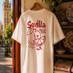 May include: A white short-sleeve t-shirt featuring a red graphic on the back. The design includes the text 'Sevilla', 'ANDALUCÍA ESPAÑA', and 'Naraajas y tapasss', alongside an illustration of an orange, a slice of orange, and a tower.
