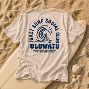 May include: A beige short-sleeve t-shirt featuring a dark blue graphic on the back. The design includes a surfer on a wave, the text BALI SURF SOCIAL CLUB ULUWATU, and the phrase GOOD WAVES, GOOD PEOPLE AND NOWHERE TO RUSH in a circular layout.
