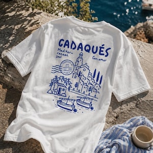 Könnte beinhalten: Ein weißes kurzärmeliges T-Shirt mit einem blauen Grafikdruck auf der Rückseite. Das Design zeigt den Text CADAQUÉS, Mediterranean soul und Con amor sowie eine Illustration einer Küstenstadt, Booten und einem Poststempel-Motiv.