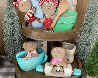 May include: A two-tiered wooden tiered tray with a black metal base. The top tier features a gingerbread person holding a milk bottle, a gingerbread person holding a wooden spoon and a green bowl, and a white ceramic house. The bottom tier features a gingerbread person in a blue coffee cup, a gingerbread person sitting on a rolling pin, and a white ceramic mug.