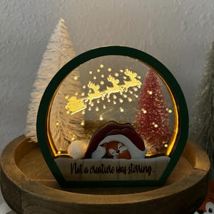 May include: A green wooden Christmas snow globe with a white and red Santa hat and a fox inside. The globe is lit up and has a gold reindeer and sleigh with snowflakes in the background. The text "Not a creature was stirring." is written on the base.