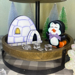 Puede incluir: Una bandeja de madera de dos niveles con temática invernal. Presenta un iglú de madera con detalles morados, un pingüino sonriente con pajarita morada y árboles de Navidad en miniatura. Cubitos de hielo transparentes esparcidos.