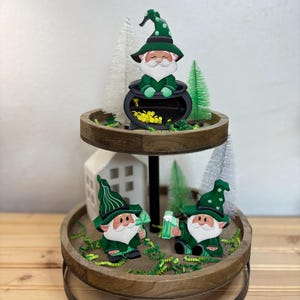 May include: Two-tiered wooden tray with a green gnome holding a pot of gold on the top tier and two green gnomes holding a beer mug and a shamrock on the bottom tier. The tray is decorated with green and white paper shreds.