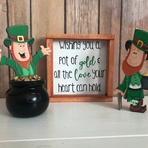 May include: Two wooden leprechaun figurines, one holding a pot of gold and the other holding a walking stick. A wooden sign reads "Wishing you a pot of gold & all the love your heart can hold."