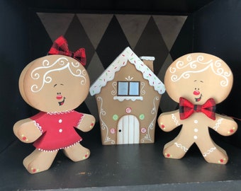 May include: A gingerbread couple stands in front of a gingerbread house. The gingerbread people are painted with white icing and have red accents. The gingerbread house is decorated with white icing and sprinkles.