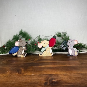 May include: Three wooden mouse figurines arranged in a row. The left mouse is grey and carries a blue bulb. The centre mouse is tan with a light blue scarf and holds a red bulb. The right mouse is grey with a light blue scarf and holds a black plug.