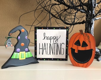 Può includere: Un cappello da strega nero con accenti verdi e viola, una zucca nera e arancione con un viso intagliato e un cartello bianco con pois neri e il testo "happy haunting" in nero.
