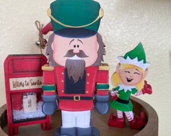 May include: A wooden nutcracker figurine with a red and gold uniform, a green hat, and a brown beard. A wooden elf figurine with a green hat, green dress, and red shoes is standing next to the nutcracker. A red wooden mailbox with the text "Letters to Santa" is in the background.